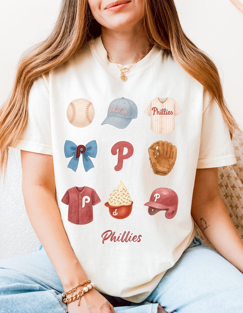 May include: Cream-colored t-shirt featuring baseball-themed illustrations. The design includes a baseball, a baseball cap, a jersey, a bow, the letter "P", a baseball glove, a shirt, a sundae, and a helmet. The word "Phillies" is written at the bottom.