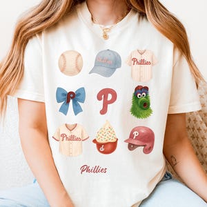May include: Cream-colored t-shirt featuring a collage of Phillies baseball-themed illustrations. The design includes a baseball, cap, jersey, bow, logo, mascot, ice cream, and helmet. The word "Phillies" is printed at the bottom.