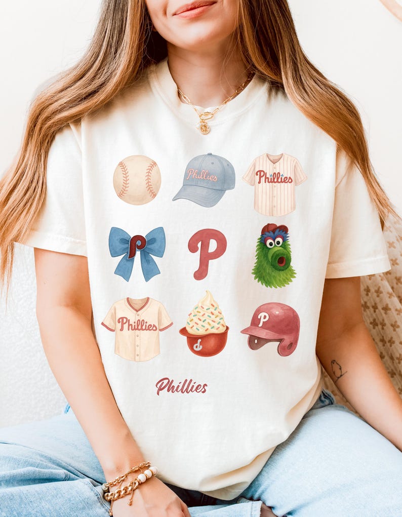 May include: Cream-colored t-shirt featuring a collage of Phillies baseball-themed illustrations. The design includes a baseball, cap, jersey, bow, logo, mascot, ice cream, and helmet. The word "Phillies" is printed at the bottom.