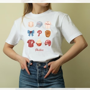 May include: White t-shirt featuring watercolor illustrations of baseball-themed items, including a baseball, cap, jersey, bow, the letter "P", glove, shirt, ice cream, and helmet. The word "Phillies" is printed below the illustrations.