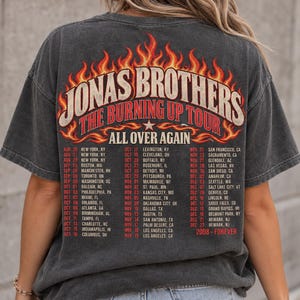 Pode incluir: Uma t-shirt de manga curta cinzento-carv&atilde;o com um gr&aacute;fico nas costas que inclui o texto JONAS BROTHERS THE BURNING UP TOUR ALL OVER AGAIN. O design apresenta chamas cor de laranja e amarelas e uma lista de datas e cidades da digress&atilde;o de 2008.