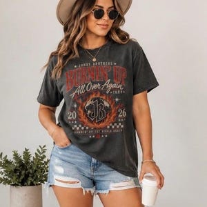 Pode incluir: Uma t-shirt cinzento-carv&atilde;o de manga curta com uma impress&atilde;o gr&aacute;fica com o texto "Jonas Brothers Burnin' Up All Over Again Tour 2026" e um log&oacute;tipo "JB" central rodeado por chamas, combinada com cal&ccedil;&otilde;es de ganga azul-claros desgastados.