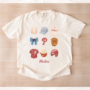 May include: Cream-colored t-shirt featuring a watercolor design of baseball-related items. The design includes a baseball, cap, jersey, bow, letter "P", glove, shirt, ice cream, and helmet. The word "Phillies" is written in red script.