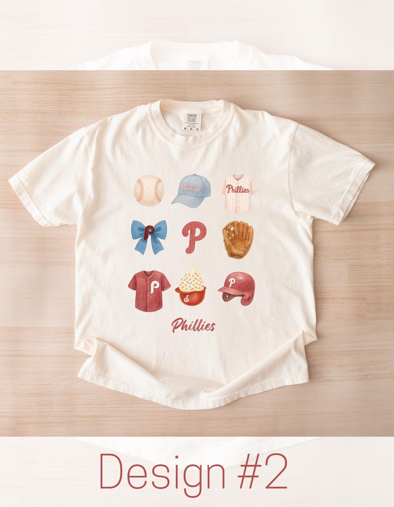 May include: Cream-colored t-shirt featuring a watercolor design of baseball-related items. The design includes a baseball, cap, jersey, bow, letter "P", glove, shirt, ice cream, and helmet. The word "Phillies" is written in red script.