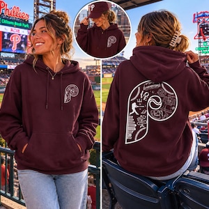 Może przedstawiać: Bordowa bluza z kapturem z białym logo Philadelphia Phillies z przodu i z tyłu. Tył zdobi duża biała litera "P" z ilustracjami o tematyce baseballowej. Bluza ma kaptur ze sznurkiem i przednią kieszeń. Pokazano również pasującą bordową czapkę z daszkiem.