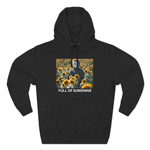 Puede incluir: Sudadera con capucha negra con un gráfico de una persona con una máscara blanca de pie en un campo de girasoles. El texto "FULL OF SUNSHINE" está impreso debajo del gráfico.