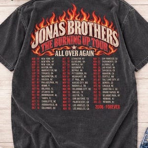 Pode incluir: Uma t-shirt cinzenta escura de manga curta com um gr&aacute;fico nas costas que apresenta chamas cor de laranja e amarelas. O texto diz Jonas Brothers The Burning Up Tour All Over Again, seguido por uma lista de datas e cidades, e 2008-Forever.