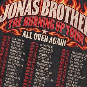 Pode incluir: Uma t-shirt cinza-carv&atilde;o com uma estampa gr&aacute;fica de chamas laranjas e amarelas ao redor do texto JONAS BROTHERS THE BURNING UP TOUR ALL OVER AGAIN. Abaixo das chamas, uma lista de datas e cidades da turn&ecirc; est&aacute; impressa em branco e vermelho.