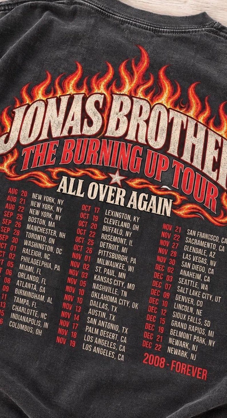 Pode incluir: Uma t-shirt cinza-carv&atilde;o com uma estampa gr&aacute;fica de chamas laranjas e amarelas ao redor do texto JONAS BROTHERS THE BURNING UP TOUR ALL OVER AGAIN. Abaixo das chamas, uma lista de datas e cidades da turn&ecirc; est&aacute; impressa em branco e vermelho.