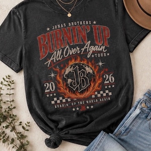 Pode incluir: Uma t-shirt cinzento-carv&atilde;o de manga curta com uma impress&atilde;o gr&aacute;fica com o texto Jonas Brothers Burnin' Up All Over Again Tour 2026. O design inclui um globo rodeado por chamas, um padr&atilde;o axadrezado e as iniciais JB no centro.