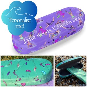 Puede incluir: Un estuche rígido para gafas de color morado con un patrón de nadadores y el texto blanco "Jane needs Vitamin Sea!", junto a un estuche turquesa con patrón de kitesurf y un paño de limpieza a juego dentro de un estuche abierto.