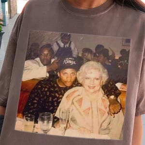 Puede incluir: Camiseta color espresso con una imagen de estilo vintage de dos personas en una mesa. La camiseta es de corte holgado y parece estar hecha de un material suave. La imagen de la camiseta tiene forma rectangular. La camiseta se lleva puesta al aire libre.