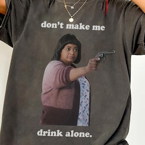 May include: Dark gray t-shirt featuring a graphic of a woman holding a gun, with the text "don't make me drink alone." The shirt has a vintage, worn look.