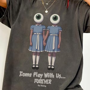 May include: Dark grey t-shirt featuring a graphic of two figures with eyeball heads, holding hands, dressed in blue dresses. The text "Come Play With Us... Forever" is printed below the graphic. The Shining.