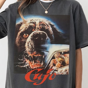 Puede incluir: Camiseta gris oscuro con un gráfico de cartel de película. El gráfico representa un perro gruñendo y la palabra "Cujo" en rojo. La camiseta tiene un corte holgado y mangas cortas.