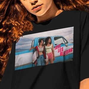 May include: Black t-shirt with a graphic of two women beside a blue truck, the words "LOVE LIES BLEEDING" are visible. The image is a colourful scene, with the text in red. The shirt is a casual wear item.