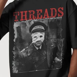 Puede incluir: Camiseta negra con la palabra "THREADS" en letras rojas en la parte superior. La camiseta presenta un gráfico en blanco y negro de una persona con uniforme y la cara vendada. La persona lleva un sombrero que dice "TRAFFIC WARDEN".