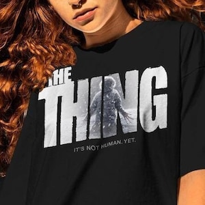 May include: Black t-shirt with a white graphic of the text "THE THING" with a silhouette of a creature behind the text. The text below the graphic reads "IT'S NOT HUMAN, YET."