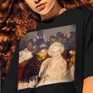 May include: Black t-shirt with a photo print of a group of people, including a woman wearing a white dress and a man wearing a black hat with the word "Kings" on it.