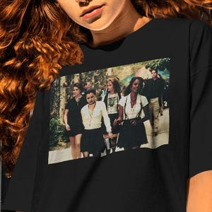 May include: A black short-sleeved t-shirt featuring a rectangular graphic print on the chest. The print displays a scene with five individuals standing outdoors, wearing white shirts and dark skirts or pants, captured in a cinematic film still style.