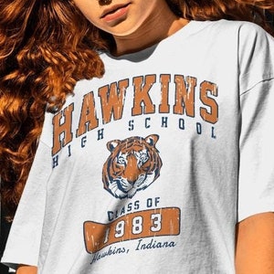 Può includere: Maglietta bianca con una grafica in stile vintage della Hawkins High School. La grafica presenta una mascotte di tigre, il testo "Hawkins High School" e il testo "Class of 1983 Hawkins, Indiana".