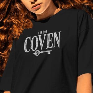 May include: A black short-sleeved t-shirt featuring a white graphic print on the chest. The design includes the text 1996 COVEN in a distressed font, positioned above a detailed illustration of an ornate skeleton key with a decorative bow.