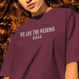 May include: A maroon short-sleeved t-shirt featuring the white text WE ARE THE WEIRDOS printed on the chest, accompanied by a small graphic of three lit candles positioned directly below the text in a centered horizontal alignment.