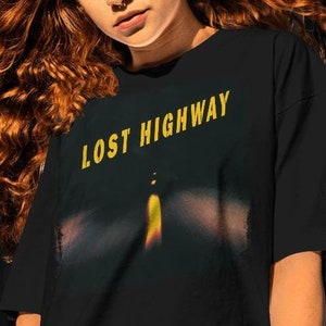 以下が含まれることがあります： 黄色の文字で「LOST HIGHWAY」と書かれた黒いTシャツ。シャツには、夜の街灯のぼやけた画像がプリントされています。