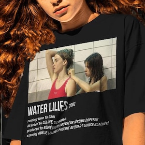 May include: A black t-shirt with a white graphic of the movie poster for "Water Lilies" (2007). The poster features two young women in a swimming pool, with the title of the film and the names of the director, producer, and actors listed below.