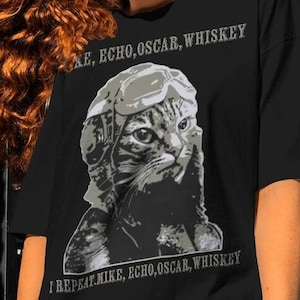 May include: A black t-shirt with a graphic of a cat wearing a pilot's helmet and goggles. The text "MIKE, ECHO, OSCAR, WHISKEY" is printed above the cat and "I REPEAT MIKE, ECHO, OSCAR, WHISKEY" is printed below the cat.