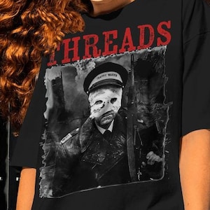 Puede incluir: Camiseta negra con un estampado gráfico de una persona con uniforme militar y una máscara. El texto "THREADS" está impreso en rojo sobre el gráfico.