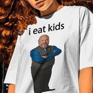 May include: White t-shirt with a graphic of a man with a blue shirt and a vest, with the text "i eat kids" above the graphic.
