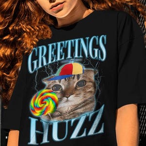 Op de afbeelding: Zwart T-shirt met een afbeelding van een kat met een pet en een regenboog lolly. De tekst "GREETINGS HUZZ" staat in een gestileerd lettertype met bliksemaccenten. De kat is het centrale element van het ontwerp.