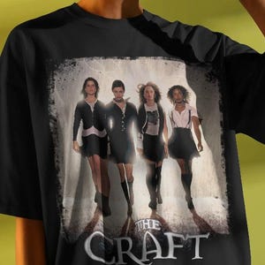 May include: Black t-shirt featuring a graphic from the movie "The Craft." The design shows four women in black and white, with the film's title in a stylized font at the bottom. The shirt is worn by a person wearing sunglasses.