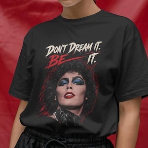 Könnte beinhalten: Ein schwarzes Kurzarm-T-Shirt mit einem Grafikdruck einer Person mit dunklem Haar, blauem Lidschatten und rotem Lippenstift. Über dem Porträt steht der Text "DON'T DREAM IT. BE IT." in weißer und roter Schrift auf schwarzem Hintergrund.