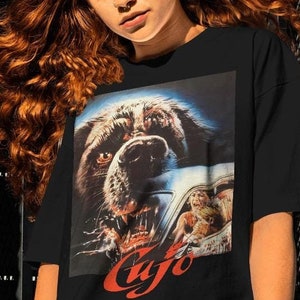 May include: Black t-shirt with a graphic print of a dog's face snarling with a car in the background. The text "Cujo" is printed in red below the image.