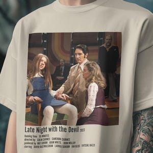 May include: A beige short-sleeved t-shirt featuring a rectangular graphic print from the film Late Night with the Devil 2023. The print shows a scene with three people in 1970s attire, with film credits and production details listed below the image.