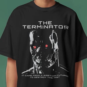 Könnte beinhalten: Ein schwarzes Kurzarm-T-Shirt mit einer Grafik eines Roboterschädels mit leuchtend roten Augen. Der Text lautet THE TERMINATOR oben und IT CAME FROM A WARTORN FUTURE TO DESTROY THE PAST unten in einer weißen, blockartigen Schriftart.