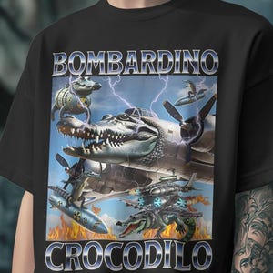 Op de afbeelding: Zwart T-shirt met een gedetailleerde afbeelding. Het ontwerp toont een vliegtuig met krokodillenthema met de tekst "BOMBARDINO CROCODILO" in een metallic lettertype. De afbeelding bevat krokodillen, vliegtuigen, bliksem en vlammen.