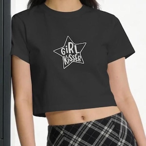 May include: A charcoal gray short-sleeved crop top featuring a white outline of a star on the chest. Inside the star, the text "GIRL KISSER" is printed in a white, hand-drawn style font. The garment is paired with a black and white plaid skirt.