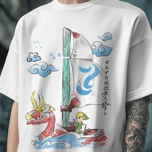 Puede incluir: Camiseta blanca con una ilustración colorida de un barco con una figura de dragón, un personaje con túnica verde y texto japonés. El diseño incluye nubes azules y elementos acuáticos.