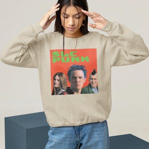 Op de afbeelding: Een beige sweatshirt met een grafische print van een band genaamd "SLC PUNK". De bandleden dragen kleurrijke kleding en hebben punk-kapsels.