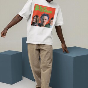 Op de afbeelding: Een wit T-shirt met een grafische print van een band genaamd "SLC PUNK". De bandleden zijn afgebeeld in de grafiek. Het shirt wordt gedragen door een persoon met een bruine huid en zwart haar.