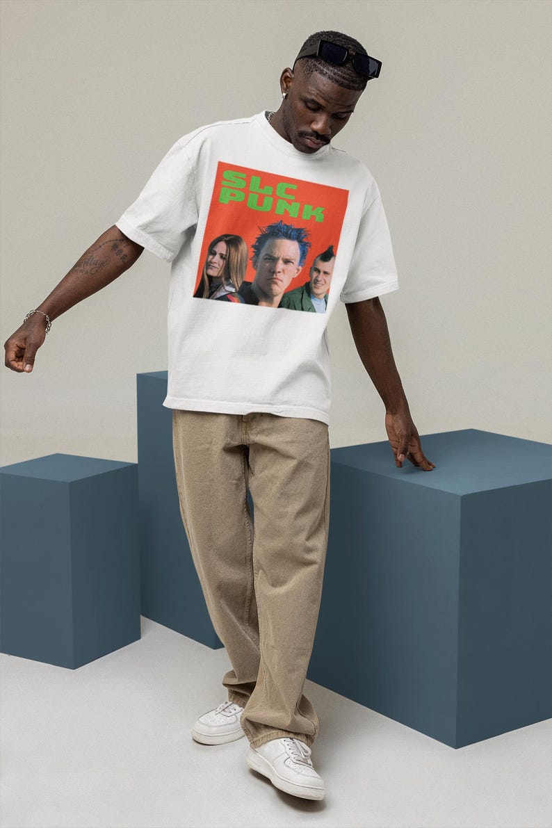 Op de afbeelding: Een wit T-shirt met een grafische print van een band genaamd "SLC PUNK". De bandleden zijn afgebeeld in de grafiek. Het shirt wordt gedragen door een persoon met een bruine huid en zwart haar.