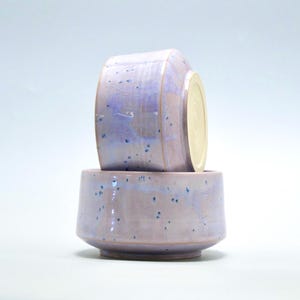 Pode incluir: Duas tigelas de cerâmica feitas à mão em um esmalte lavanda com salpicos azuis. Uma tigela é empilhada sobre a outra, mostrando os detalhes internos e externos. As tigelas têm uma forma geométrica única e um acabamento liso.