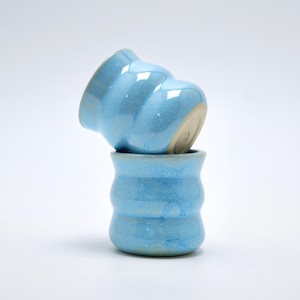 Può includere: Due tazze in ceramica impilate con una smaltatura lucida color azzurro. Le tazze presentano una distinta silhouette ondulata e scanalata con tre creste orizzontali. La tazza inferiore è dritta, mentre quella superiore è inclinata sul bordo.