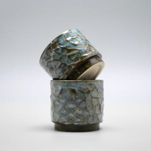 Op de afbeelding: Twee keramische bekers met een getextureerde, blauwgroene glazuur. De bekers zijn gestapeld, waarbij de ene op de andere rust. De bekers hebben een uniek, organisch patroon en een donkerbruine basis.