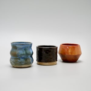 Peut inclure: Trois tasses en céramique faites à la main. La tasse de gauche est bleue avec un motif ondulé. La tasse du milieu est marron foncé avec une surface texturée. La tasse de droite est orange et marron.