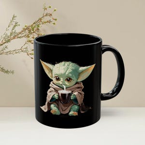 Puede incluir: Taza de cer&aacute;mica negra con una ilustraci&oacute;n de dibujos animados de un personaje alien&iacute;gena verde bebiendo de una taza marr&oacute;n oscuro. El alien&iacute;gena lleva una t&uacute;nica marr&oacute;n y tiene orejas grandes. La taza tiene un asa negra.