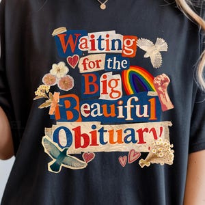 May include: Black t-shirt with the text "Waiting for the Big Beautiful Obituary" in a colorful, collage-style design. The shirt features a rainbow, flowers, and other decorative elements. The shirt is a comfortable, casual wear.